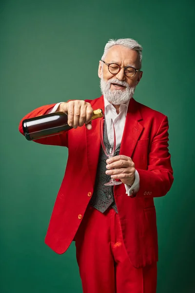 Alegre Santa con barba blanca vertiendo copa de flauta con champán sobre fondo verde, concepto de invierno - foto de stock