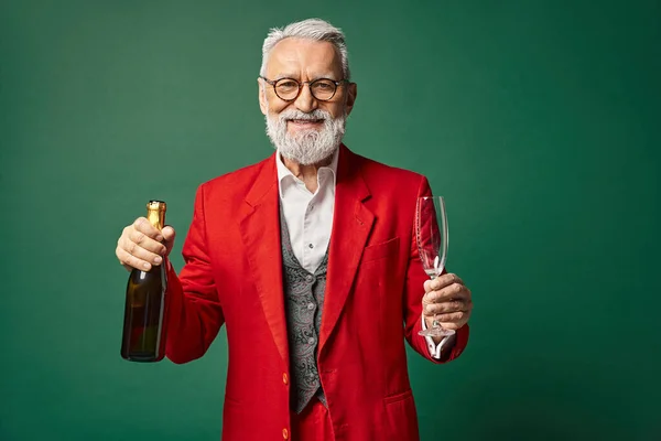 Весела Санта з білою бородою позує з шампанським та флейтовим склом на зеленому тлі, зимова концепція — стокове фото