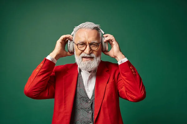 Homem elegante vestido como Papai Noel usando fones de ouvido no fundo verde escuro, conceito de Natal — Fotografia de Stock