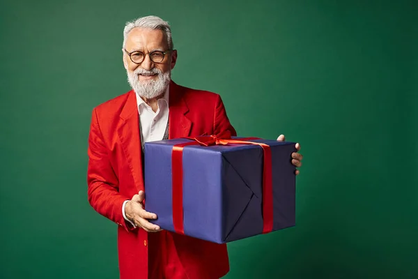 Elegante Papai Noel com barba e óculos segurando enorme presente e sorrindo sinceramente, conceito de inverno — Fotografia de Stock