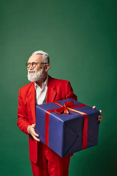 Stylish classy Santa holding huge present on green backdrop and looking away, winter concept — Stock Photo