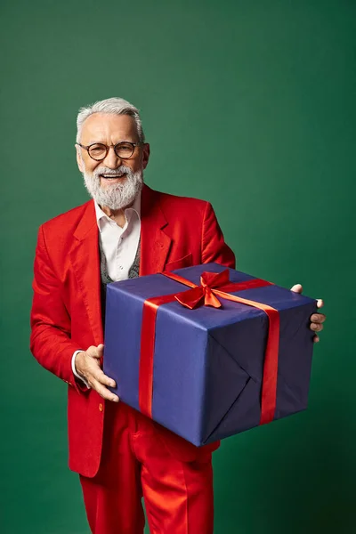 Веселий Санта в стильному червоному вбранні тримає величезний подарунок і посміхається на камеру, зимова концепція — стокове фото