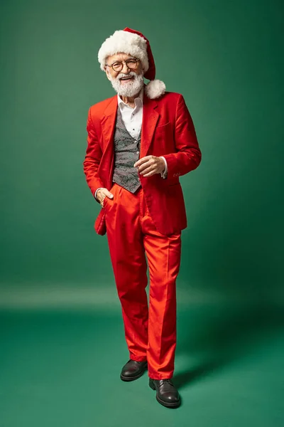 Hombre alegre vestido como Santa con sombrero navideño posando con una mano en el bolsillo, concepto de invierno - foto de stock