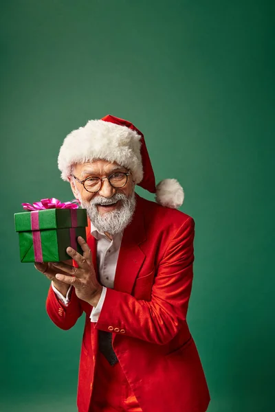 Beau Père Noël élégant en chapeau rouge et tenue élégante montrant présent à la caméra, concept d'hiver — Photo de stock
