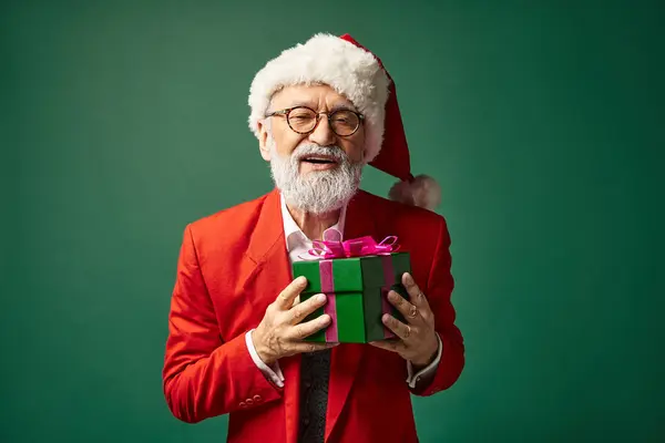 Hombre elegante feliz vestido como Santa con sombrero rojo sosteniendo presente mirando a la cámara, concepto de invierno - foto de stock