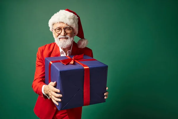 Hombre elegante guapo en sombrero rojo navideño y gafas que sostienen regalo azul enorme, concepto de invierno - foto de stock