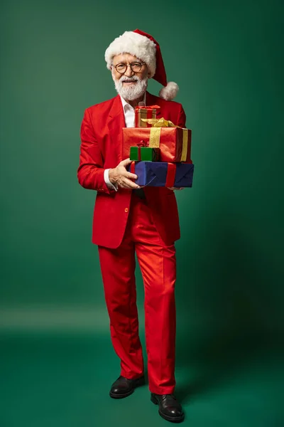 Elegante Santa em vermelho elegante traje segurando pilha de presentes em pano de fundo verde, conceito de inverno — Fotografia de Stock