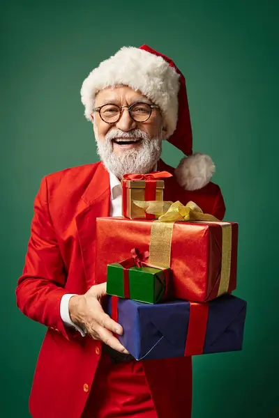 Retrato de Papá Noel elegante en traje rojo sosteniendo montón de regalos sonriendo a la cámara, concepto de invierno - foto de stock