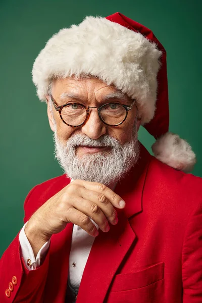 Elegante Santa con sombrero rojo y gafas mirando a la cámara con la mano bajo la barbilla, concepto de invierno - foto de stock