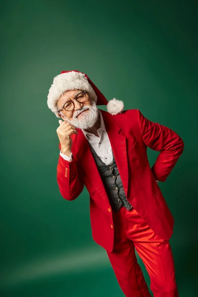 Allegro uomo elegante vestito da Babbo Natale in occhiali e cappello in posa su sfondo verde scuro, concetto invernale — Foto stock