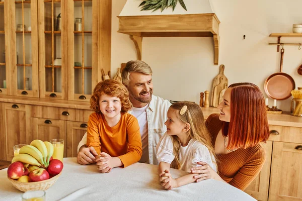 Padres sonrientes con hija e hijo sentados cerca de frutas frescas y jugo de naranja en la acogedora cocina - foto de stock