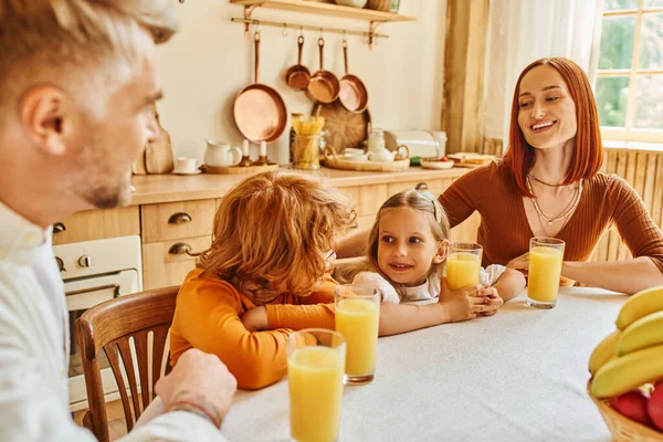 Padres alegres con hija e hijo cerca de zumo de naranja y frutas durante el desayuno en la cocina - foto de stock