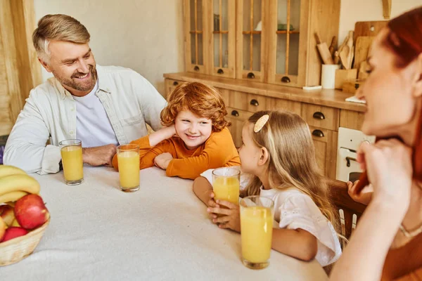 Padres alegres con hija e hijo cerca de zumo de naranja fresco y frutas durante el desayuno en la cocina - foto de stock