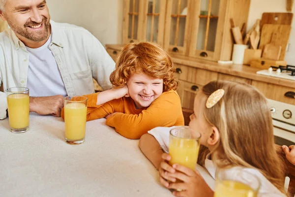 Irmãos despreocupados sorrindo um para o outro perto de suco de laranja fresco e pais na cozinha em casa — Fotografia de Stock