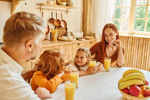 Alegre hermano sonriendo el uno al otro cerca de jugo de naranja fresco y los padres en la cocina en casa - foto de stock