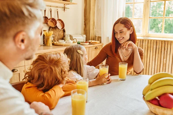 Улыбающаяся женщина смотрит на детей возле свежего апельсинового сока и во время завтрака на кухне дома — стоковое фото