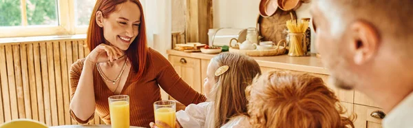 Joyful woman looking at children near fresh orange juice during breakfast in cozy kitchen, banner — Stock Photo