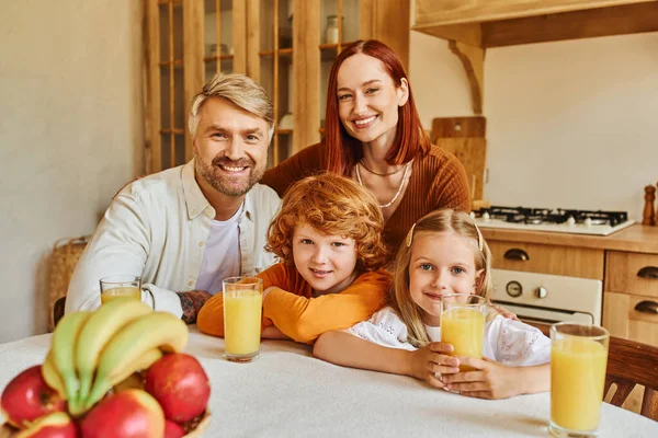 Casal alegre com crianças adoráveis olhando para a câmera perto de frutas frescas e suco de laranja na cozinha — Fotografia de Stock