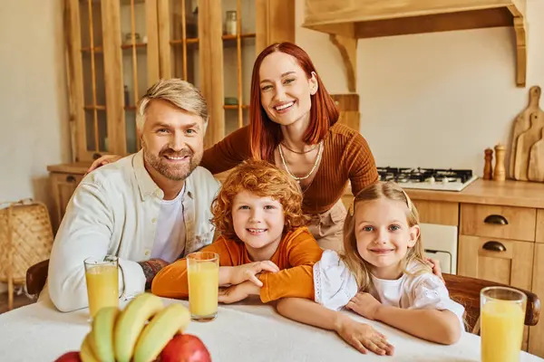 Joyous parents with adorable kids looking at camera near fresh fruits and orange juice in kitchen — Stock Photo