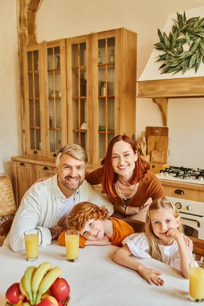 Parents avec des enfants mignons regardant la caméra près pendant le petit déjeuner dans la cuisine confortable, visages souriants — Photo de stock