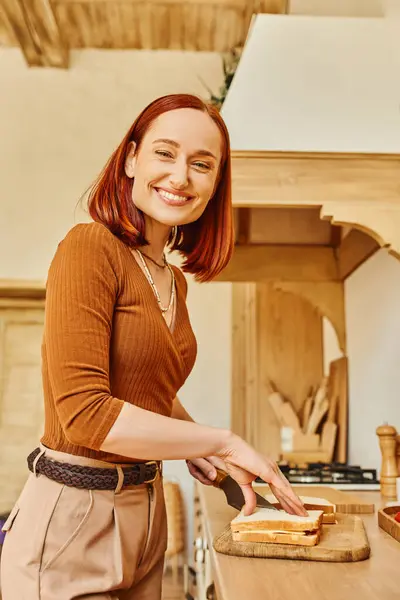 Joyful redhead woman looking at camera and preparing delicious sandwiches for breakfast in kitchen — Stock Photo