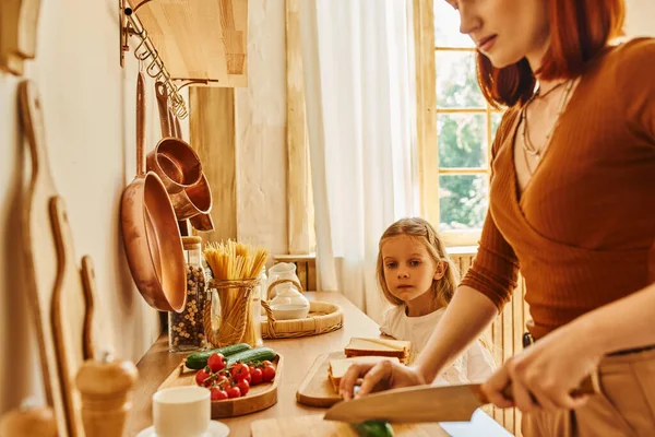 Sorridente madre tagliare le verdure preparare la colazione vicino figlia con panino sul bancone della cucina — Foto stock