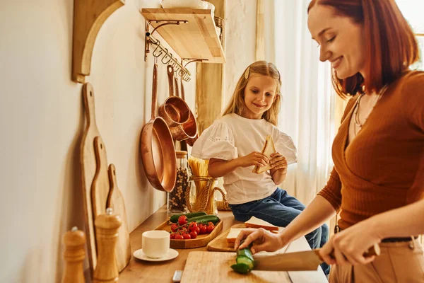Mulher feliz cortando legumes preparando café da manhã perto da filha com sanduíche no balcão da cozinha — Fotografia de Stock