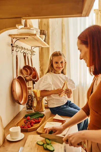 Menina feliz sentada com sanduíche no balcão da cozinha perto de mãe sorridente preparando café da manhã — Fotografia de Stock