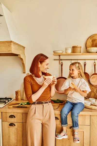 Веселые мать и дочь едят вкусные бутерброды на завтрак и в современной уютной кухне — стоковое фото
