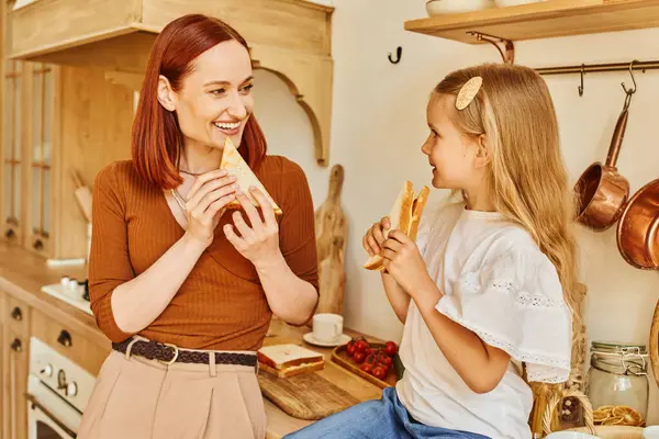 Веселые мать и дочь едят вкусные бутерброды на завтрак и в современной уютной кухне — стоковое фото