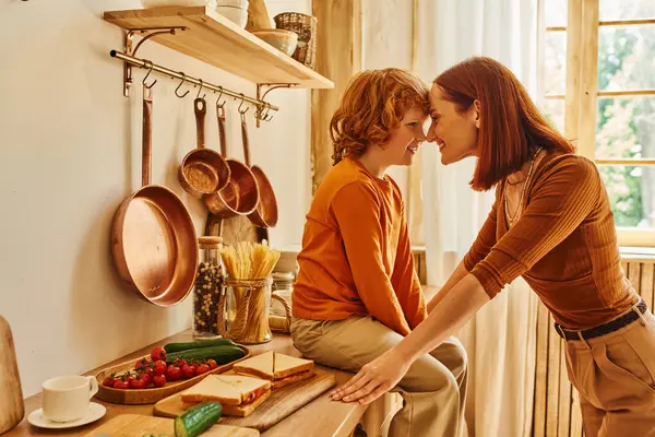 Criança alegre no balcão da cozinha perto de sanduíches e legumes frescos cara a cara com mãe sorridente — Fotografia de Stock