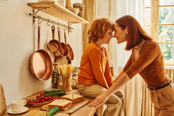 Joyful kid on kitchen counter near sandwiches and fresh vegetables face to face with smiling mother — Stock Photo
