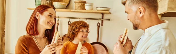Fröhliche Eltern und niedlicher Sohn essen Sandwiches zum Frühstück in der modernen Küche, horizontales Banner — Stockfoto