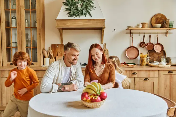 Playful kids running around happy parents sitting with smartphone near fresh fruits in cozy kitchen — Stock Photo