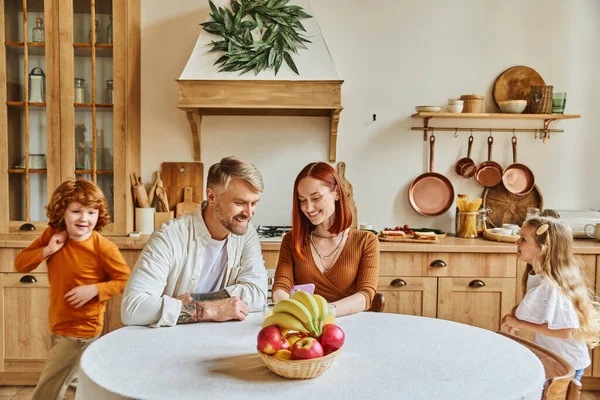 Fröhliche Kinder herumlaufen glückliche Eltern sitzen mit Smartphone neben frischem Obst in der gemütlichen Küche — Stockfoto