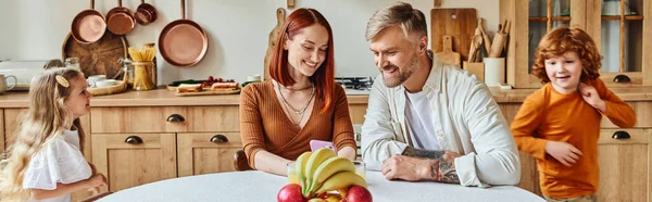 Cheerful kids running around parents sitting with smartphone near fresh fruits in kitchen, banner — Stock Photo