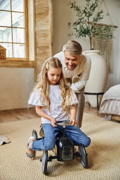 Smiling man assisting cute daughter riding toy car in modern living room at home, playing together — Stock Photo