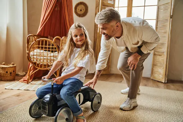 Cheerful man assisting cute daughter riding toy car in modern living room at home, playing together — Stock Photo