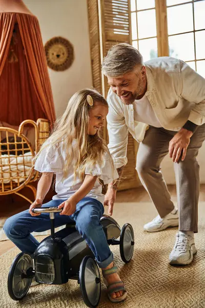 Заботливый отец помогает счастливой дочери верхом на игрушечной машине в уютной гостиной дома, играть вместе — стоковое фото