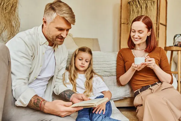 Lächelnde Frau mit Kaffeetasse in der Nähe von Vater und Tochter beim Lesen von Büchern im Schlafzimmer, gemeinsames Lernen — Stockfoto
