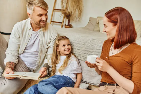 Joyful woman with coffee cup near father and daughter reading book in bedroom, learning together — Stock Photo