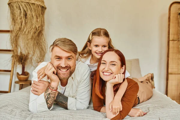 Fröhliche Eltern mit unbeschwerter Tochter, die es sich auf dem Bett bequem macht und in die Kamera schaut, gemütliches Wohnumfeld — Stockfoto