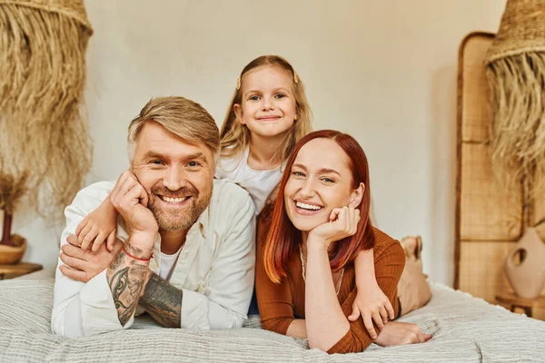 Happy tattooed man with smiling wife and adorable daughter looking at camera on bed at home — Stock Photo