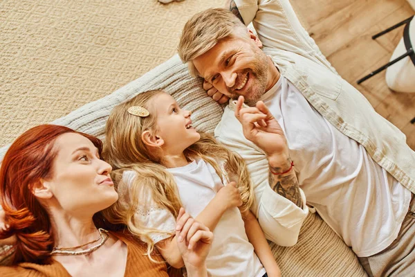 Top view of cheerful parents with cute daughter lying down and talking on bed, fun and laughter — Stock Photo