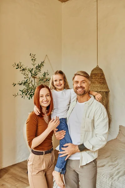 Cheerful husband and wife holding cute daughter and looking at camera in cozy bedroom, love and care — Stock Photo