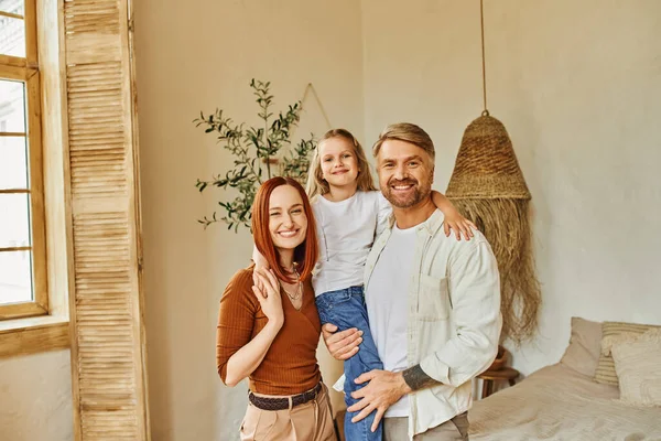 Joyful husband and wife holding cute daughter and looking at camera in cozy bedroom, love and care — Stock Photo