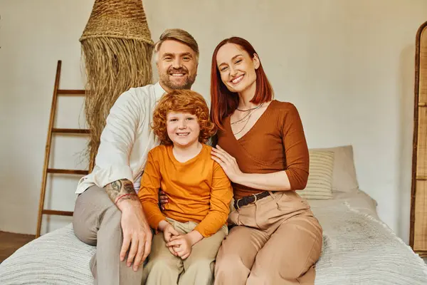 Cheerful parents with redhead son sitting on bed and looking at camera in cozy home environment — Stock Photo