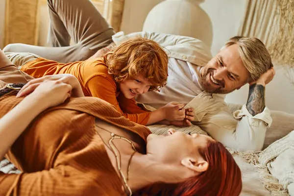 Feliz tatuado homem olhando para esposa e ruiva filho se divertindo na cama em casa, tempo de relaxamento — Fotografia de Stock