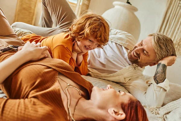 Smiling tattooed man looking at wife and redhead son having fun on bed at home, relaxation time — Stock Photo