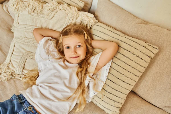 Menina loira feliz deitado em travesseiros macios com as mãos atrás da cabeça e olhando para a câmera no quarto acolhedor — Fotografia de Stock